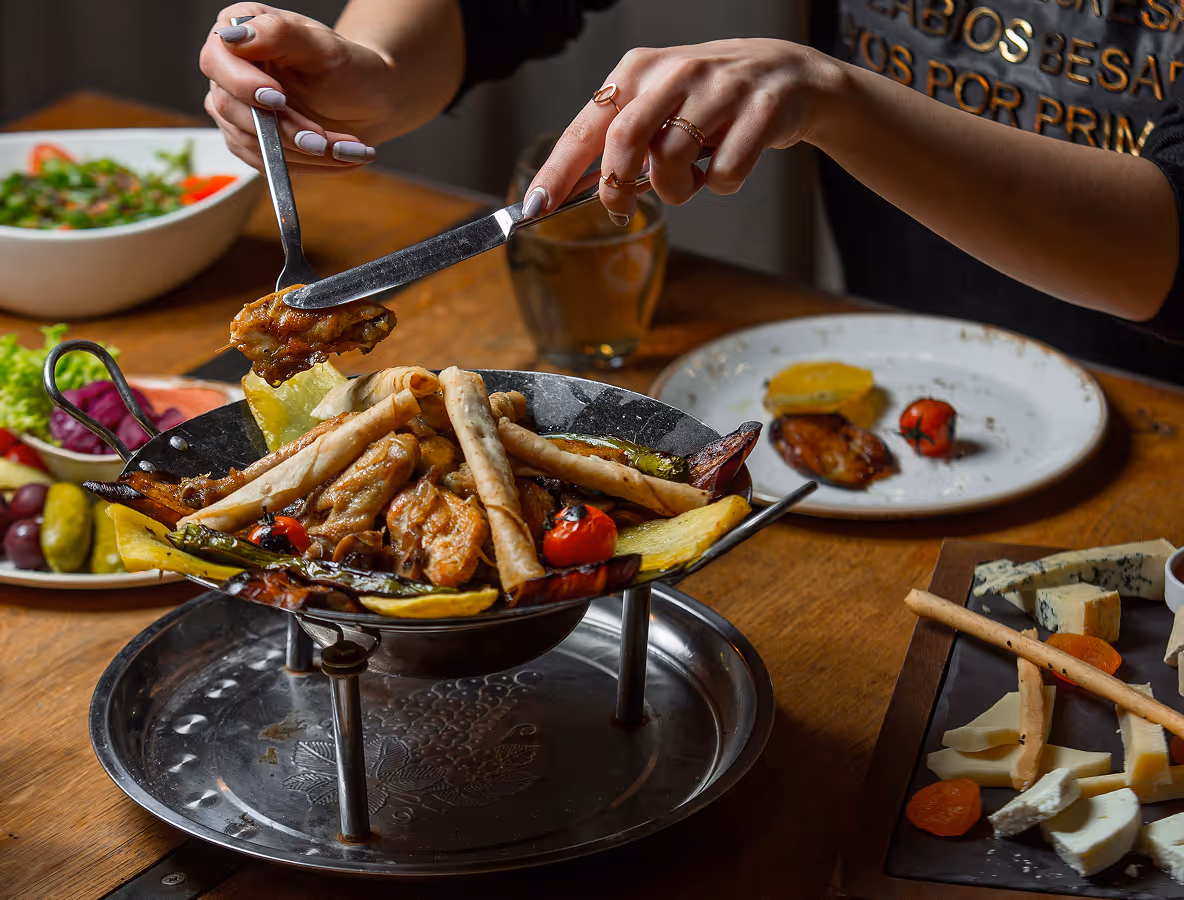 Hands using knife and fork to serve grilled chicken and vegetables from a metal serving dish on a wooden table with salad and cheese plates nearby.